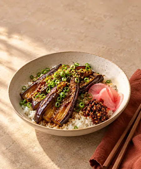 A glossy miso-glazed eggplant rice bowl with scallions, sesame, pickled radish, and chili crisp on a warm neutral tabletop.