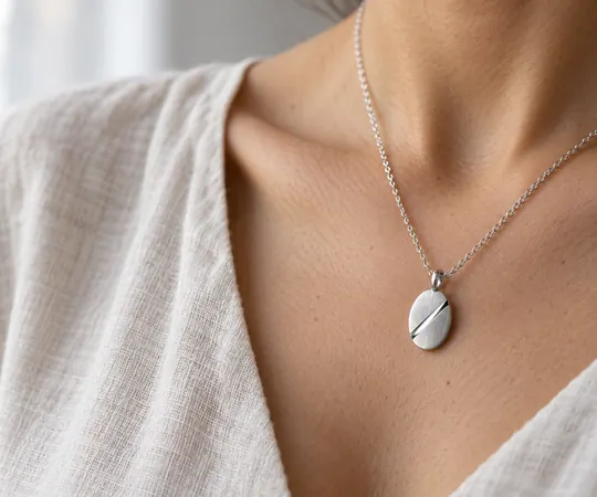 A brushed oval silver pendant with a polished diagonal groove rests against a collarbone above an ivory linen neckline.