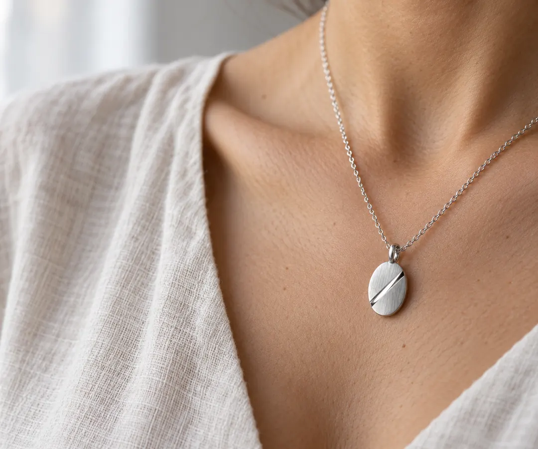 A brushed oval silver pendant with a polished diagonal groove rests against a collarbone above an ivory linen neckline.