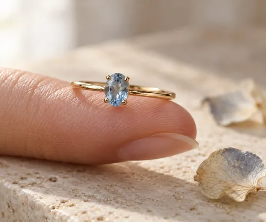 A petite gold ring with a pale-blue gemstone rests on a fingertip in a softly lit ivory product scene.